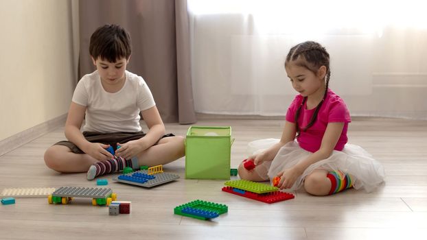 Two children, a boy and a girl, are sitting on the floor in the room, play in multi-colored plastic construction set made of blocks. Wide baner