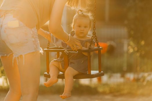 Toddler baby girl on a swing on the warm summer evening. Mother is swinging her young daughter on a sunny playground.