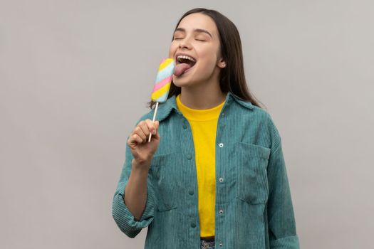 Portrait of positive woman with dark hair licking multicolored ice cream, feels hungry, tasting dessert, wearing casual style jacket. Indoor studio shot isolated on gray background.