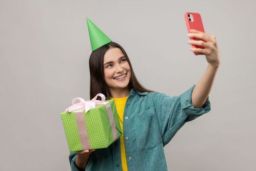Portrait of smiling woman holding present box in hands and taking selfie or having video call with client, wearing casual style jacket. Indoor studio shot isolated on gray background.