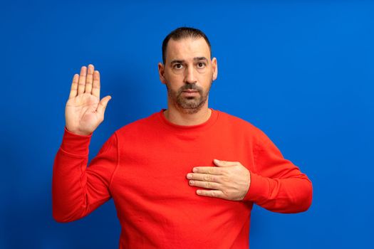Photo of serious and confident bearded hispanic man with hand on chest promising oath isolated over blue colored background
