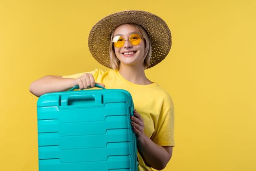 Young pretty woman with carry-on suitcase on yellow background. Teenager traveling with blue luggage bag for airplane hand baggage. Summer travel, vacation concept. High quality photo