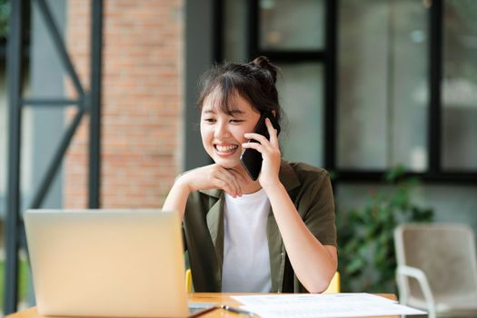 Young asian businesswoman uses a mobile phone to contact a customer, working hard at the office using laptop data graphs, planning for improvement, analyzing and strategizing for business growth. Business concept