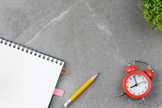 Open notepad with a red pen, an alarm clock and a flower in a pot on a gray marble background. Top view.