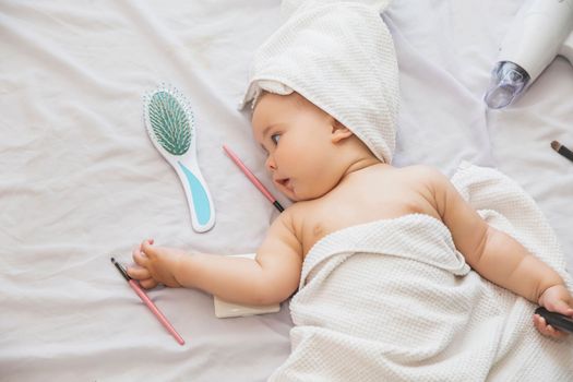 charming baby with a white towel on her head lies near the hairdryer, cosmetics and comb