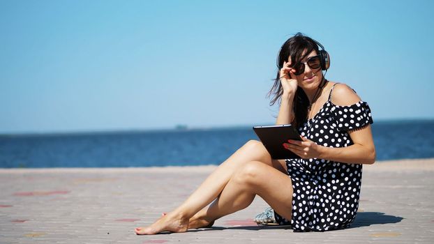 beautiful brunette, woman in sun glasses, and headphones, works on a tablet, listening to music , sitting on the beach, barefoot, against the sea. wind waves her hair. High quality photo