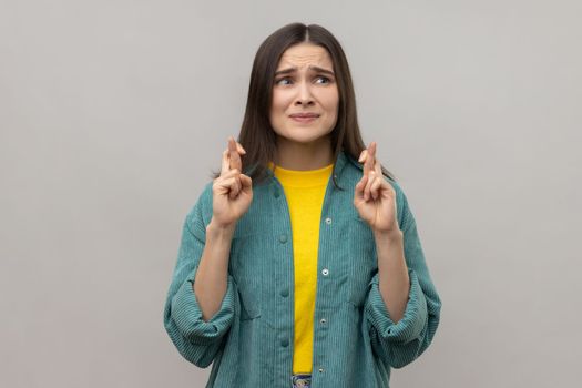 Portrait of woman standing with crossed fingers for good luck, heartily making wish, praying and hoping for miracle, wearing casual style jacket. Indoor studio shot isolated on gray background.