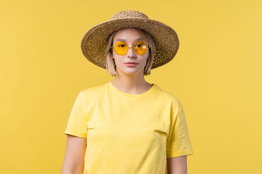 Portrait of young pretty blonde woman on summer yellow studio background. Confident sunny outfit with sunglasses and straw hat. High quality photo
