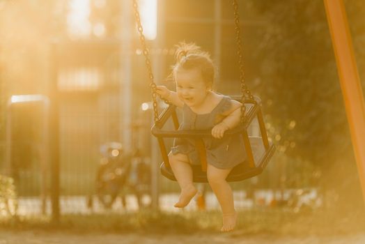 Toddler baby girl on a swing on the warm summer evening. Mother is swinging her young daughter on a sunny playground.