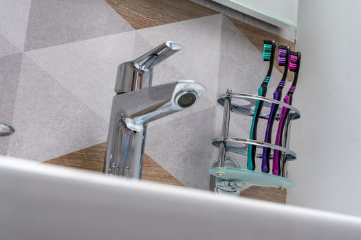multi-colored toothbrushes stand on a special stand in the bathroom interior. a sink and a modern mixer are visible. products for daily oral hygiene