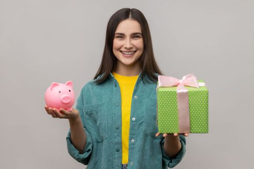 Portrait of happy woman holding wrapped gift box and piggy bank, save money, advertising, looking smiling at camera, wearing casual style jacket. Indoor studio shot isolated on gray background.