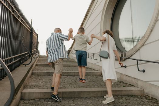 A photo from the back of a family is climbing the stairs of the palace in an old European town. A happy father, mother, and son are holding hands in the evening.