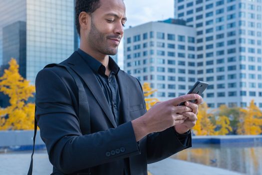 Businessman using mobile phone app texting outside of office in urban city with skyscrapers buildings in the background. Young black man holding smartphone for business work. High quality photo