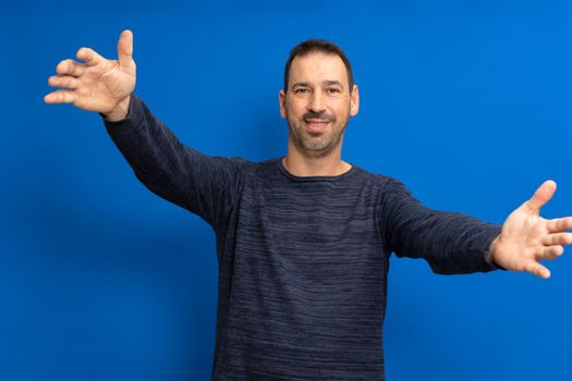Happy handsome bearded hispanic man standing with arms raised, wants to hug his friend, isolated on blue background... I love you. I miss you body language