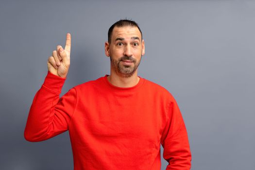 Portrait of a bearded hispanic man dressed in red pullover happy excited pointing finger at copyspace isolated on gray background