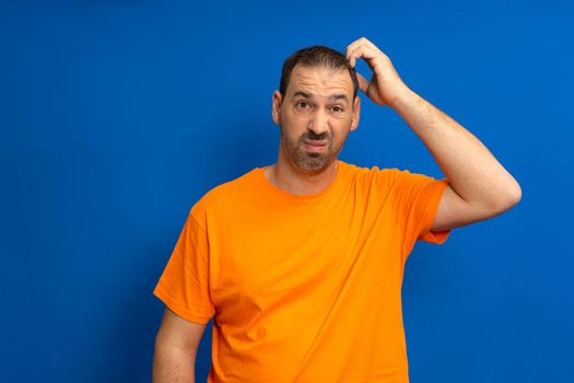 Miserable and gloomy guy with messy hair, frowning and looking unsatisfied while scratching head, standing over blue background. Something heavy fell on his head, man feels pain.