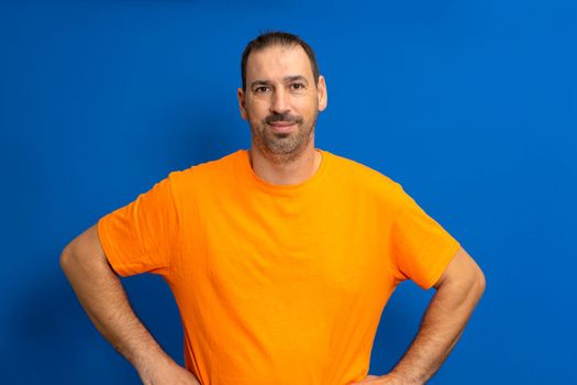 Cheerful bearded man in orange t-shirt posing isolated on blue background, studio portrait. People lifestyle concept. Mock up copy space. Standing with arms akimbo on waist blinking