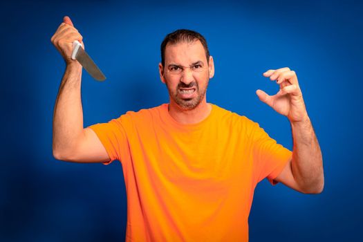 Hispanic man with a beard wearing an orange t-shirt in an aggressive attitude wielding a knife ready to attack, isolated on a blue studio background