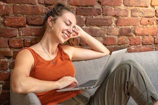 Happy blonde woman with laptop sitting on couch against brick wall. Remote work. High quality photo