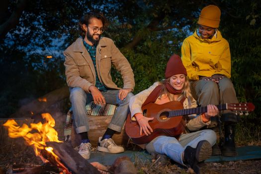Three happy diverse friends having fun playing music and enjoying bonfire camping in nature at night. Group of people chilling at fire in the evening, camping in the forest near lake. High quality photo