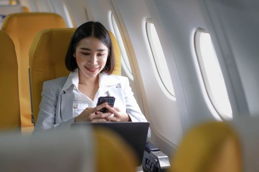 Asian woman passenger sitting in airplane near window and reading news from social networks or using travel applications in smartphone.