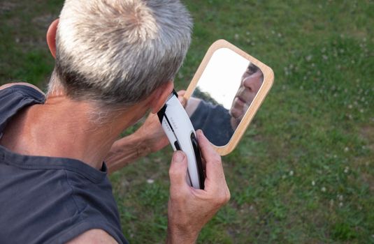 a gray-haired middle-aged man shaves his hair with a clipper in a garden on a green lawn and looks into a table mirror. High quality photo