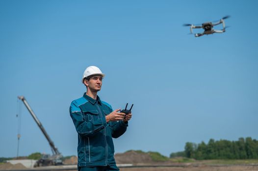 A man in a helmet and overalls controls a drone at a construction site. The builder carries out technical oversight