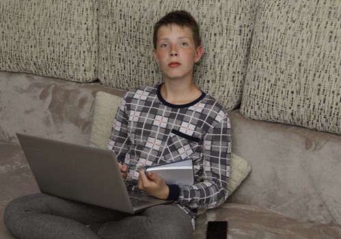 Pensive boy is engaged in distance learning..One boy is sitting on the couch with a laptop.