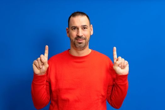 Portrait of a happy hispanic bearded man pointing fingers up at copyspace isolated over blue background