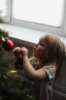 Little girl decorating christmas tree with toys and baubles. Cute kid preparing home for xmas celebration