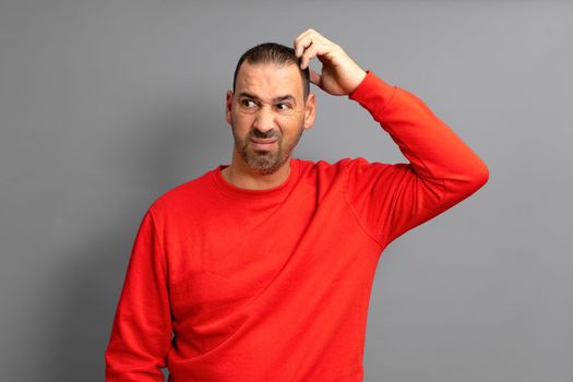 Bearded Hispanic man in red sweater frowning and looking dissatisfied while scratching his head, standing over blue background.