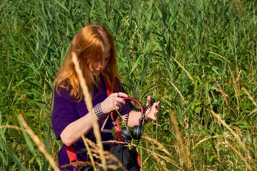 A female person who has reached puberty but has not yet married. Pretty red haired girl puts on headphones among tall green grass.