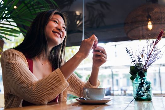 smiling asian woman sitting in a coffee shop opening the sugar sachet for her coffee, concept of relaxation and leisure