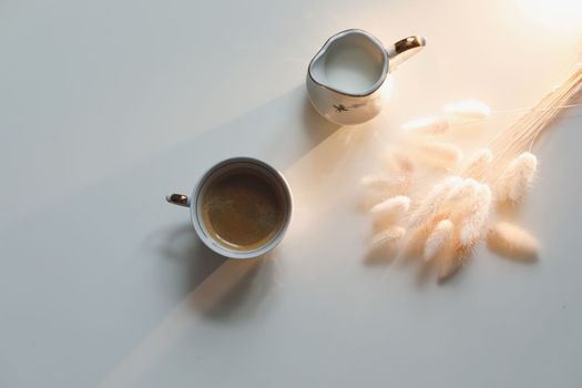 White cup with coffee and milk jug on a white wooden background, closeup. Energy breakfast, morning routine concept. coffee cup and milk pitcher on white table background