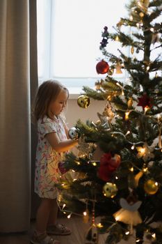 Little girl decorating christmas tree with toys and baubles. Cute kid preparing home for xmas celebration