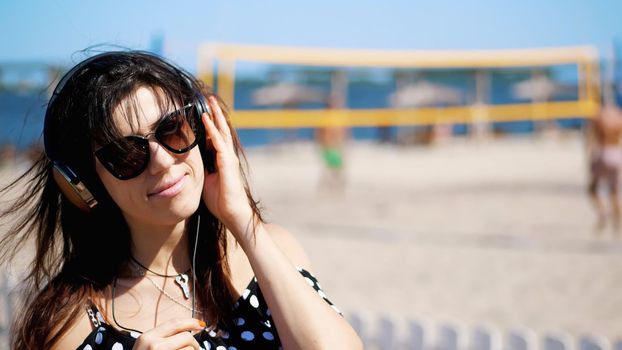 portrait, beautiful girl in sun glasses wearing big black headphones, listening to music from smartphone, on beach, on hot summer day, against background of beach volleyball. High quality photo