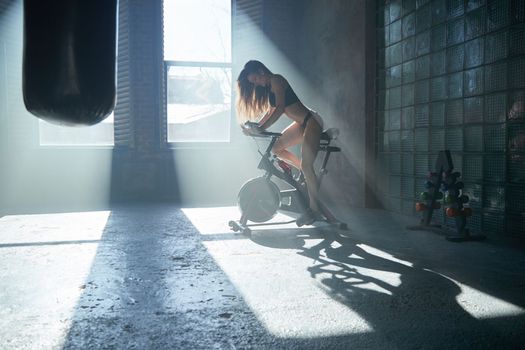 The beautiful fitness girl with a sport body twists bicycle pedals in gym, large loft-style room, beautiful light from windows, sun beams, the stationary exercise machine the bicycle. High quality photo