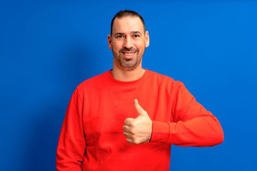 Bearded latin man wearing a red sweater raising his thumb up in approval while smiling at the camera in a friendly attitude isolated over blue background