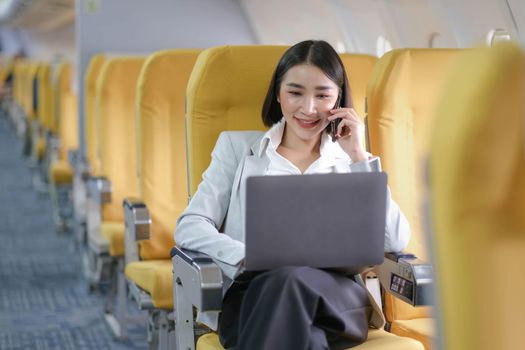 Asian woman passenger sitting in airplane near window and reading news from social networks or using travel applications in smartphone.