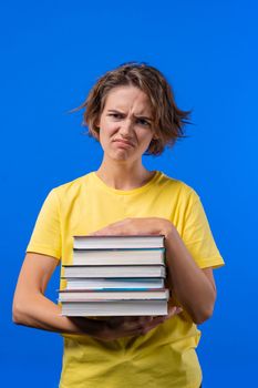 Lazy woman student is dissatisfied with amount of books homework on blue background. Girl with short hair in displeasure, she is annoyed, discouraged frustrated by studies. High quality photo