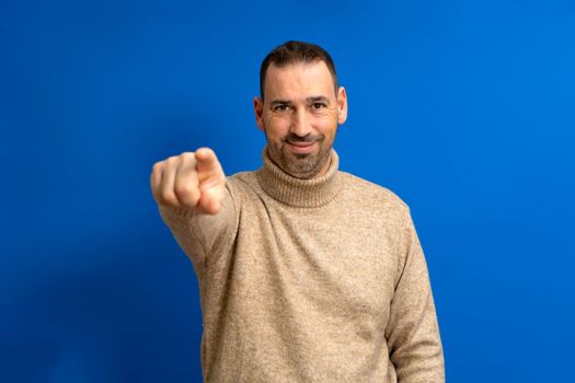 Portrait of a smiling bearded hispanic man in brown turtleneck pointing finger at camera isolated over blue background