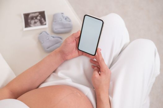 Faceless pregnant woman sits on a white sofa and holds a smartphone. Fetal ultrasonic screening
