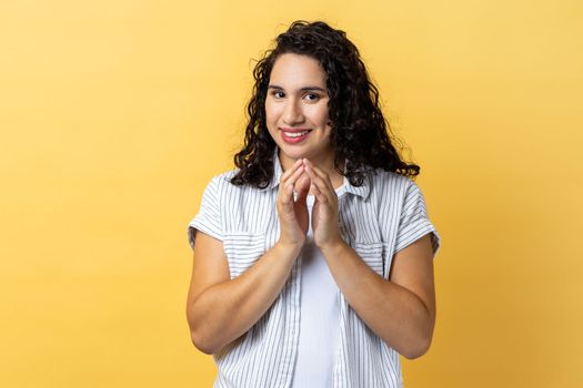 Portrait of attractive devious cunning woman with dark wavy hair clasping hands and smirking mysteriously, scheming cheats, evil prank. Indoor studio shot isolated on yellow background.