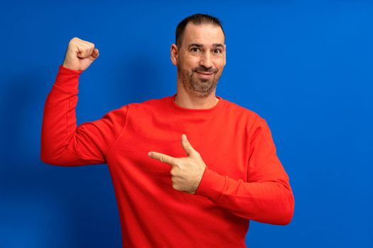 Handsome funny bearded latino man pointing at his biceps and smiling over blue background.