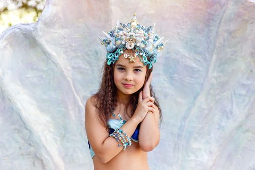 A beautiful little girl in a turquoise crown and mermaid costume is standing outdoors, in a large seashell. Close up. Copy space