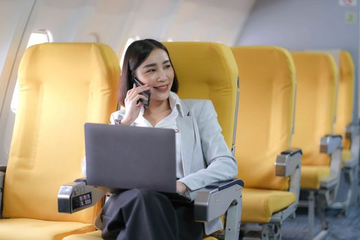 Asian woman passenger sitting in airplane near window and reading news from social networks or using travel applications in smartphone.