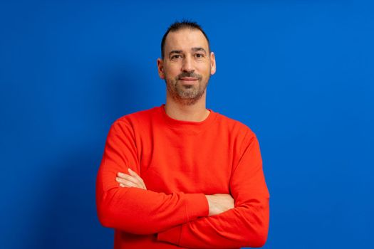 Portrait of smart cheerful attractive bearded hispanic man with arms crossed wearing bright red pullover isolated over blue colored background