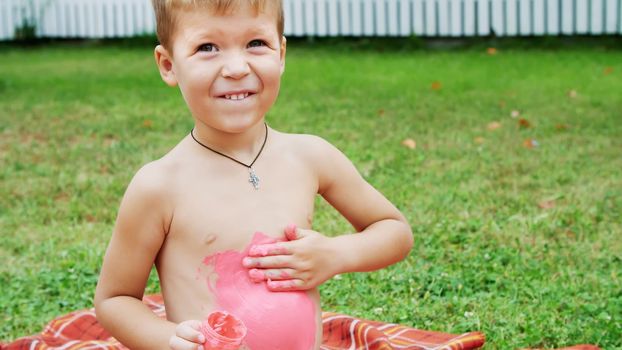 a little child, a four-year-old boy playing, painting with finger paints, decorating himself, in the garden, sitting on a blanket, on grass, lawn, in the summer. he's having fun. High quality photo