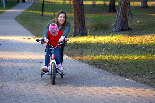 show or explain to someone how to do something. Cheerful mom teaches her child to ride a bike in autumn spring park.