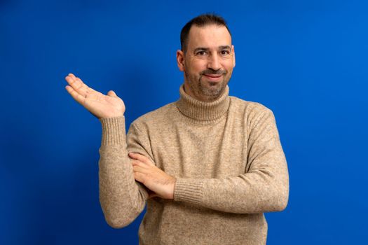Hispanic man with beard and brown turtleneck sweater in skeptical attitude offering something in the palm of his hand, isolated on blue background.
